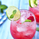close up of two glasses with pink drink ice cubes and slices of lime on a blue table